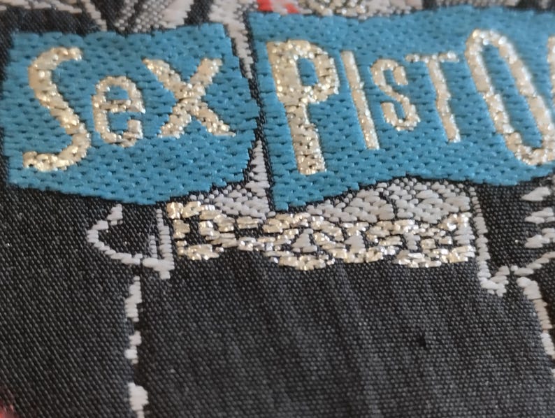 May include: Close-up of a textile with the words "Sex Pistols" in gold lettering on a turquoise blue background. The background is a dark, patterned fabric with silver and white accents. The overall aesthetic suggests a punk rock theme.