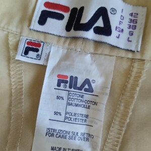 Puede incluir: Primer plano de una etiqueta de prenda beige con el logotipo de FILA en azul marino, rojo y blanco. La etiqueta incluye informaci&oacute;n sobre tallas y detalles de la composici&oacute;n de la tela: 50% algod&oacute;n y 50% poli&eacute;ster. Tambi&eacute;n se ve el texto "MADE IN TURKEY".
