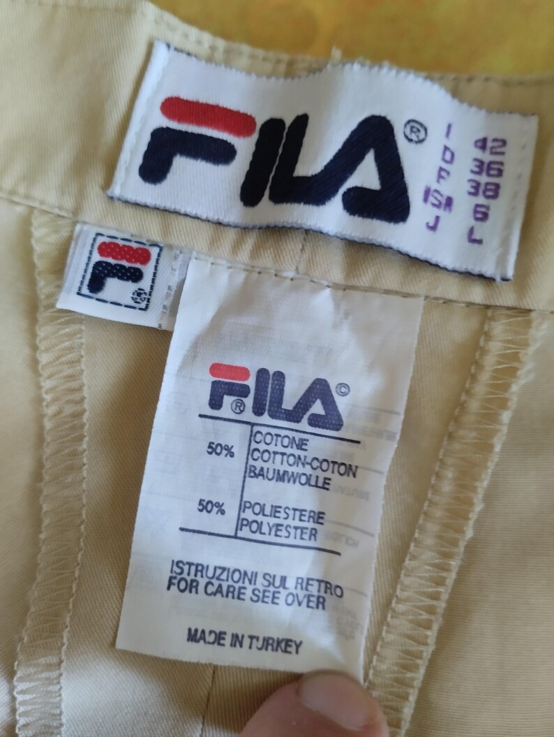 Puede incluir: Primer plano de una etiqueta de prenda beige con el logotipo de FILA en azul marino, rojo y blanco. La etiqueta incluye informaci&oacute;n sobre tallas y detalles de la composici&oacute;n de la tela: 50% algod&oacute;n y 50% poli&eacute;ster. Tambi&eacute;n se ve el texto "MADE IN TURKEY".