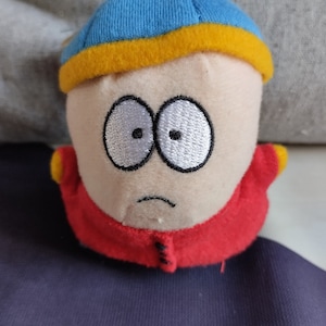 Puede incluir: Un juguete de peluche de un personaje de dibujos animados que lleva un gorro azul y amarillo y una chaqueta roja. El personaje tiene una expresión triste y ojos blancos.