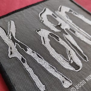 Könnte beinhalten: Ein grau gestickter Aufnäher mit dem Bandnamen "Korn" in weißer, stilisierter Schrift. Der Aufnäher hat einen schwarzen Rand und den Text "©2000 Korn U.L.T. G" am unteren Rand. Der Aufnäher besteht aus einem strukturierten Stoff.