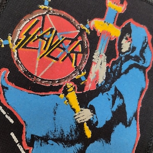 Peut inclure: Écusson carré noir avec le nom du groupe "SLAYER" en rouge et blanc. Une figure en robe bleue tenant une épée et un bâton jaune est représentée. Le texte "UNDER LICENSE TO BROCKUM" est imprimé au bas de l'écusson.