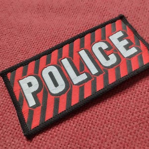Może przedstawiać: Prostokątna naszywka z czarnym obszyciem, na której widnieje słowo POLICE napisane pogrubionymi białymi wielkimi literami. Tło składa się ze wzoru w ukośne czerwone i czarne paski, tworząc kontrastowy wygląd tego przedmiotu identyfikacyjnego.