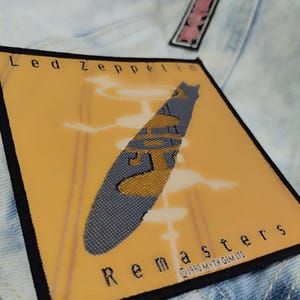 Puede incluir: Un parche bordado amarillo y gris con el texto "Led Zeppelin Remasters" y una imagen estilizada de un dirigible Zeppelin.