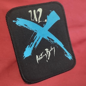 Puede incluir: Un parche rectangular de tela negra con un borde cosido. El diseño presenta el texto blanco U2 en la parte superior, una gran X azul con estilo de pincelada en el centro y el texto blanco Achtung Baby en letra cursiva en la parte inferior.