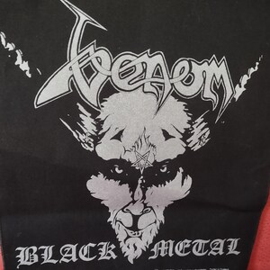 Può includere: Toppa in tessuto nero con il nome della band "Venom" in argento, sopra un'illustrazione argentata di un volto demoniaco con un pentagramma. Le parole "BLACK METAL" sono stampate in lettere gotiche argentate.