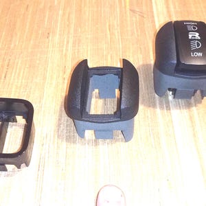 May include: Three black and gray plastic automotive switch components. One piece features a black top with white text reading HIGH, a headlight icon, the letter R, another headlight icon, and LOW. The other two pieces are hollow rectangular housings.