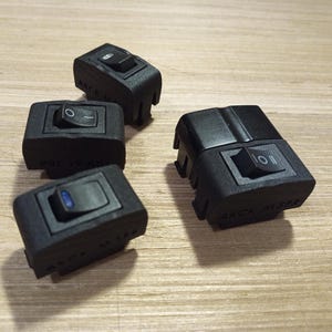 Op de afbeelding: Vier zwarte rechthoekige elektrische tuimelschakelaars met getextureerde behuizingen. Drie schakelaars hebben kleine tuimelknoppen met gedrukte symbolen zoals O, I en II. Eén schakelaar heeft een blauw indicatielampje. De tekst AKCK is zichtbaar op de basis.