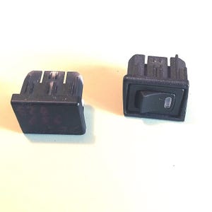 May include: Two black rectangular rocker switches with a matte finish. One switch is positioned to show the front face with a small rectangular indicator light, while the other is angled to display the side profile and the mounting clips.