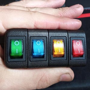 以下が含まれることがあります： 4つの照明付きロッカースイッチを備えた黒いコントロールパネル。 スイッチは緑、青、黄色、赤です。 各スイッチには白いインジケーターライトが付いています。