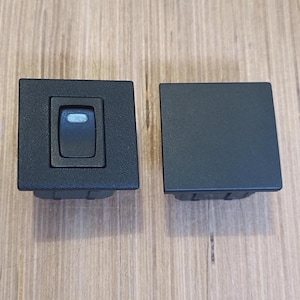 May include: Two black electrical rocker switches. One switch has a rectangular button with a white indicator light. The other switch is a solid black square. Both switches have a rectangular base.