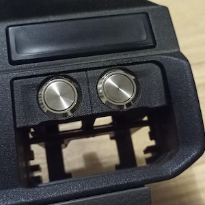 Può includere: Primo piano di un pannello in plastica nera con due pulsanti argentati. I pulsanti sono rotondi e hanno una finitura liscia e metallica.