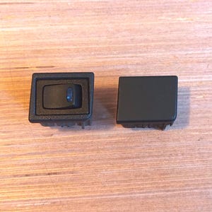 May include: Two black automotive switches on a wooden surface. One switch has a rocker, and the other is a blank cover. The switches are rectangular with a textured surface.