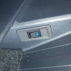 Puede incluir: Un interruptor basculante negro con una luz indicadora azul, encastrado en un marco rectangular gris oscuro. El interruptor está montado en una superficie texturizada gris oscuro, posiblemente en el interior de un vehículo.