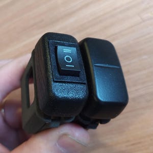 Op de afbeelding: Twee zwarte schakelaars die samen zijn gemonteerd. De schakelaar links heeft een zwarte knop met een wit "I/O" label. De schakelaar rechts is een lege schakelaar.