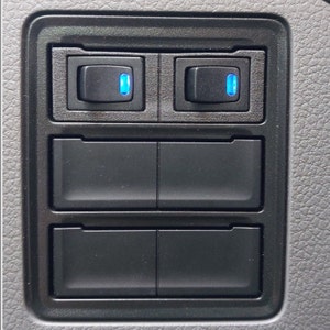 May include: A black panel with four rectangular buttons. The top two buttons have blue LED lights. The bottom two buttons are blank.