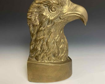 May include: A brass eagle head sculpture, featuring detailed feather textures and a strong beak. The sculpture is mounted on a rectangular base, showcasing a classic design. The eagle's gaze is focused forward, conveying a sense of strength and vigilance.