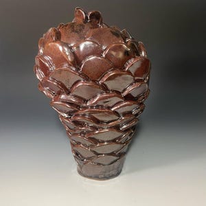 May include: A handcrafted, brown ceramic vase with a unique, textured surface. The vase features a layered, scale-like design, creating a visually interesting and organic form. The vase is approximately 12 inches tall.