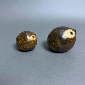 May include: Two rounded ceramic sculptures with a dark brown, mottled glaze. Each piece features a single, prominent eye design with a cream-colored almond-shaped outline and a black center, resembling a stylized bird or creature figure.