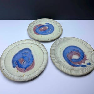May include: Three round ceramic plates with a speckled off-white base glaze. Each plate features a central hand-painted design consisting of a swirling blue spiral layered over a muted mauve circular patch, with concentric rings along the rim.