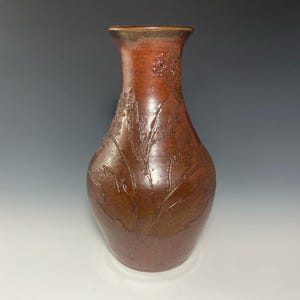 May include: A brown ceramic vase with a textured surface and a floral design. The vase has a narrow neck and a wide base.