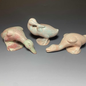 May include: Three ceramic duck figurines in shades of pink, green, and blue. The ducks are in various poses, with one preening, one looking down, and one with its head extended. The figurines have a speckled texture.
