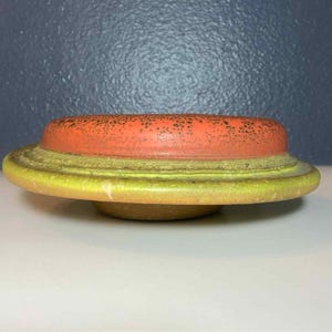 May include: A ceramic disc golf disc with a flat, rounded shape. The top is orange with dark speckles, and the rim is yellow-green. The disc is designed for flight and is used in the sport of disc golf.