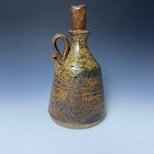 May include: Handmade ceramic bottle with a cork stopper. The bottle features a handle and a textured surface with brown, yellow, and green hues. The design has a rustic, artisanal aesthetic, suitable for decorative or functional use.