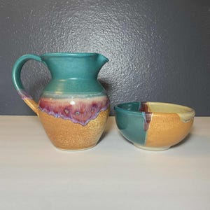 May include: A handmade ceramic pitcher and bowl set. The pitcher is teal at the top, transitioning to a purple and gold base. The bowl is split, with teal and gold sections. Both pieces have a textured finish.