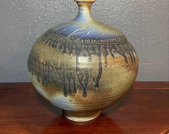 May include: A handcrafted ceramic vase with a rounded body and a narrow neck. The vase features a blend of earthy tones, including blue, brown, and green, with a dripped glaze effect. The vase is approximately 10 inches tall.