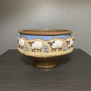 May include: A ceramic bowl with a brown rim and base, featuring a light blue band with white sheep and blue floral accents. The bowl has a wide opening and a pedestal base, suitable for decorative use.