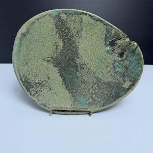 May include: An oval-shaped ceramic plate with a textured, mottled surface in shades of sage green, dark charcoal, and teal. The plate features a prominent, irregular hole near the right edge and rests on a thin, gold-toned metal display stand.
