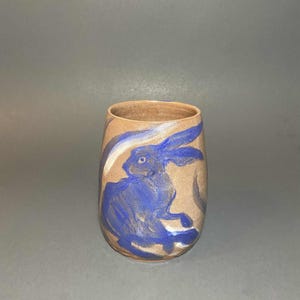 May include: A tan ceramic tumbler with a blue rabbit design. The rabbit is painted in a stylized, illustrative manner. The tumbler has a smooth, rounded shape and a slightly flared rim.