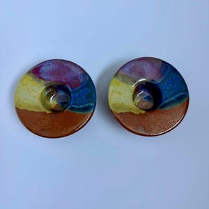 May include: Two circular ceramic candle holders featuring a multi-colored glaze. The surface displays bands of earthy terracotta, vibrant yellow, deep blue, and magenta purple, with a smooth, glossy finish and a central indentation for a candle.