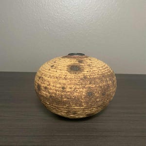 May include: A round, handcrafted ceramic vase with a textured surface. The vase is primarily tan with dark brown spots and a dark brown opening. The vase is sitting on a dark brown surface.