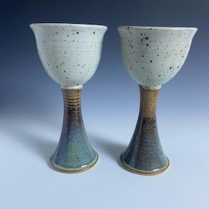 May include: Two handmade ceramic goblets. The cups are white with brown speckles. The stems transition from brown to blue-green. The base is a light brown. The goblets are displayed against a gradient blue background.