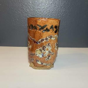 May include: A handmade ceramic tumbler with a textured, brown glaze. The cylindrical cup features a decorative pattern of dark brown and iridescent accents. The base is a lighter, unglazed clay, and the interior is not visible.