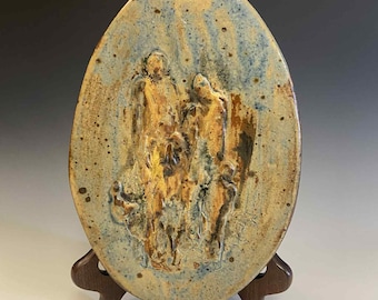 May include: Oval ceramic wall art with a relief sculpture of figures. The artwork features a blue and tan color palette with a speckled finish. The piece is displayed on a wooden easel, showcasing the textured surface and artistic detail.