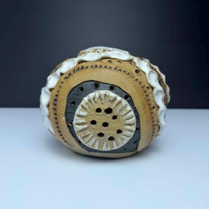 May include: A handcrafted ceramic sphere with a textured surface. The sphere features a tan base with a black and white circular design. The top edge has a white, ruffled trim. The design includes small holes and carved details.