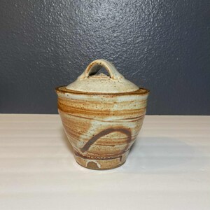 May include: A handmade ceramic jar with a lid. The jar is a light beige color with brown swirls and a handle on the lid. The jar is approximately 5 inches tall and 4 inches wide.