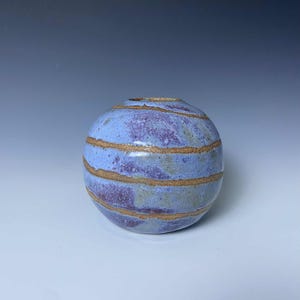 May include: A small, round ceramic vase with a light blue and purple color scheme. The vase features horizontal bands of gold-colored glaze. The handmade pottery has a unique, organic shape and texture, suitable for home decor.
