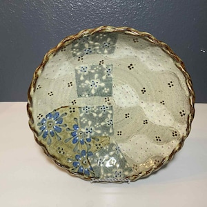 May include: A ceramic plate with a scalloped edge and a patchwork design. The plate features a mix of colors, including blue, green, and brown. The design includes floral and dotted patterns. The plate is displayed on a metal stand.