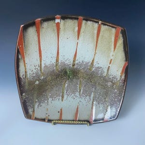 May include: A square ceramic plate with rounded corners featuring a crackle glaze finish. The surface displays vertical orange stripes over a cream-colored base, with a textured, speckled brown and green band across the center of the plate.