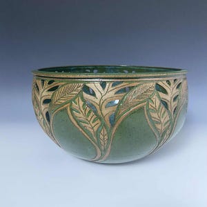 May include: A handcrafted ceramic bowl with a green glaze and leaf cutouts. The bowl features a light brown rim and detailed leaf patterns carved into the surface. The bowl is a decorative piece.