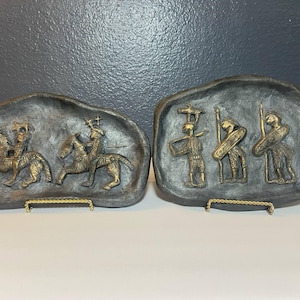 May include: Two bronze-colored relief sculptures depicting figures on horseback and warriors with spears and shields. The sculptures are mounted on dark gray, irregularly shaped plaques, displayed on decorative gold-toned stands.
