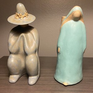May include: Two ceramic figurines, one wearing a wide-brimmed hat with floral accents and a blue and brown robe, the other wearing a light blue robe with a brown strap.