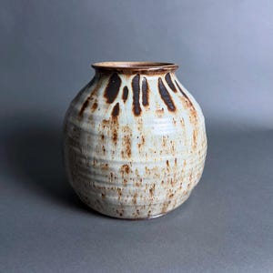May include: A round ceramic vase with a narrow neck and a wide, bulbous body. The surface features a cream-colored glaze with vertical brown drip patterns near the rim and scattered brown speckles across the textured, horizontally ribbed exterior.