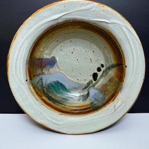 May include: A round ceramic bowl with a wide, textured off-white rim. The interior features a glossy glaze with blended earthy tones of brown, green, and blue, accented by dark speckles and a series of small black dots near the center basin.
