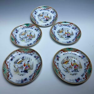May include: Five small, round porcelain plates with a gold rim. Each plate features a detailed, colorful scene of a person, birds, and a pagoda. The plates have a decorative border with red, green, and blue accents.