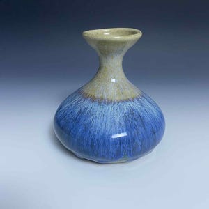 May include: A handmade ceramic vase with a rounded base and a flared neck. The vase features a gradient of colors, transitioning from a light tan at the top to a deep blue at the base. The blue has a streaked effect, adding visual interest.
